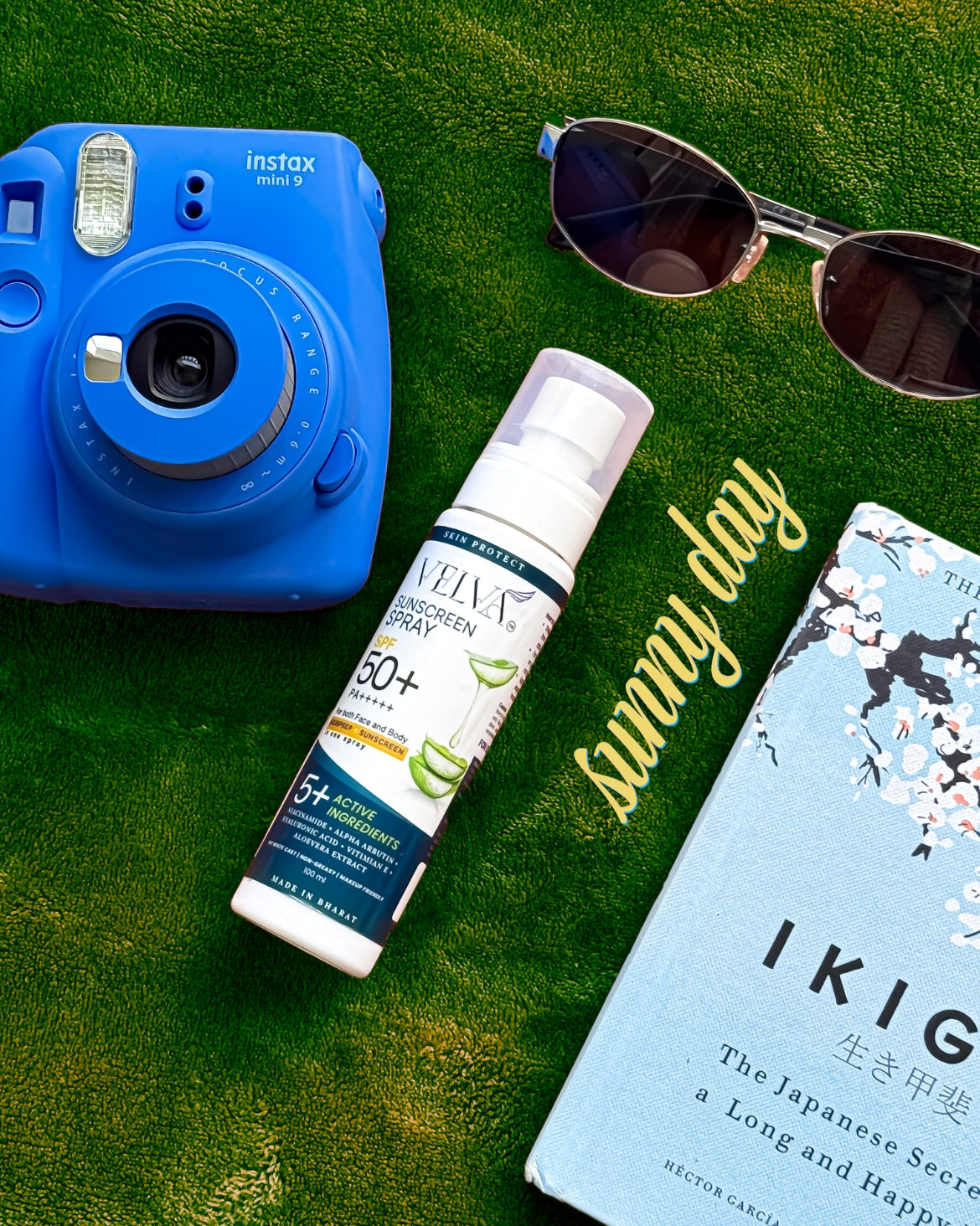 Blue Instax camera, sunscreen bottle, sunglasses, and book on grass